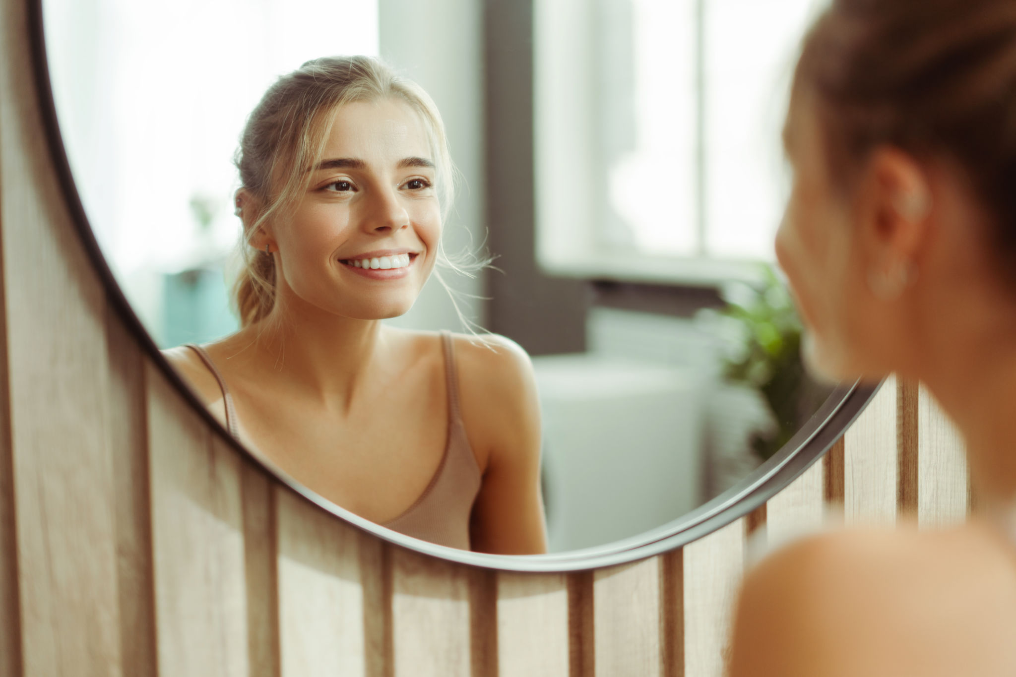 Smiling, confident woman with make up wearing tank top, looking in mirror, standing in bathroom Smiling, confident woman with make up wearing tank top, looking in mirror, standing in bathroom