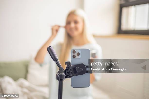 young beautiful blonde woman films a makeup lesson on her phone camera. pretty lady hosts a make-up tutorial stream - content creation stock pictures, royalty-free photos & images