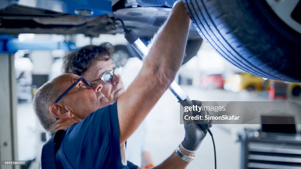 Male Mechanic And Manager Discussing Repairs Underneath Car At Workshop