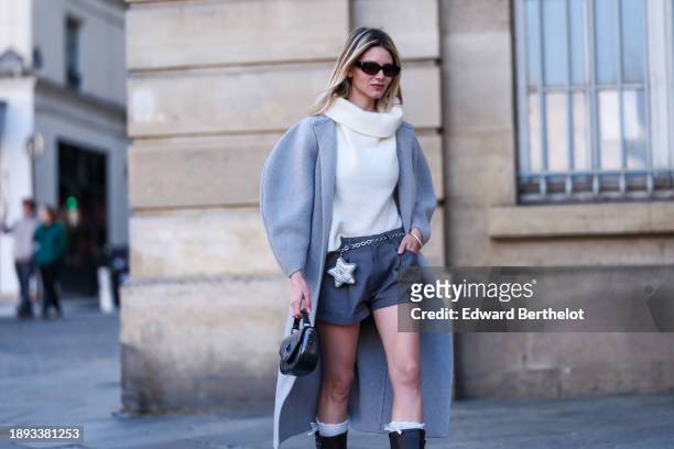 Natalia Verza wears sunglasses, gray mini shorts by Frankie shop, a white large turtleneck wool pullover by Sezane, a Chanel star-shaped belt bag, a...