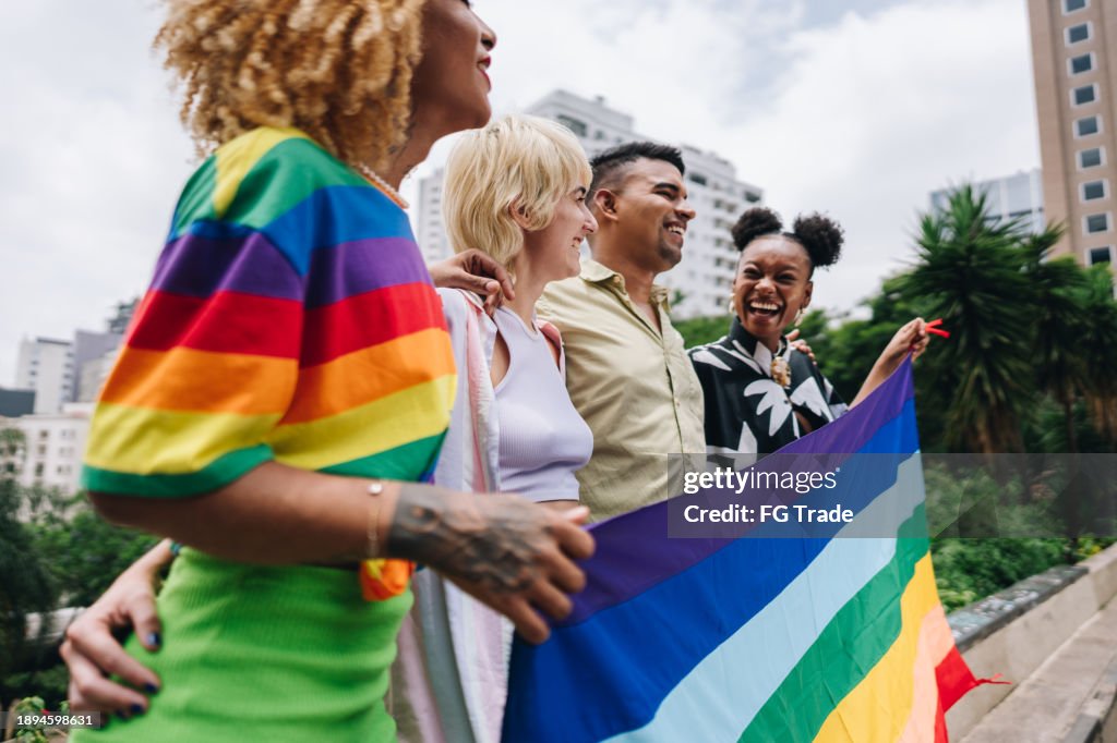 Junge LGBTQIA+-Freunde unterhalten sich und halten eine Regenbogenflagge im Freien