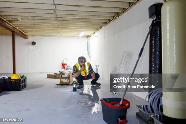 puzzled mature inspector in safety vest, searching for the cause of the leak in the flooded basement - kelder stockfoto's en -beelden