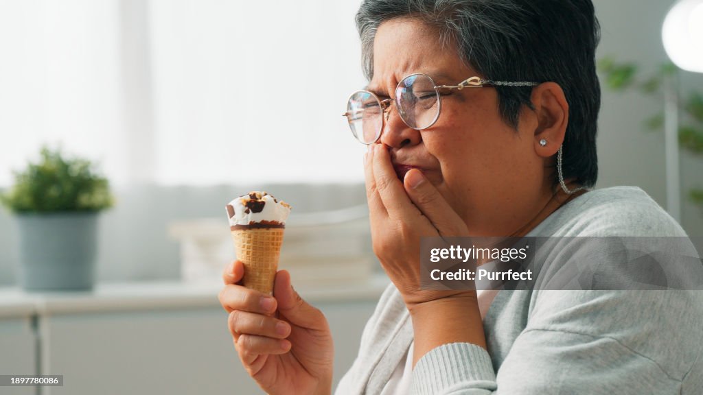 Elderly Asian grandmother suffering from sensitive teeth while eating ice cream and sitting on couch at home, Senior woman suffering from dental problem, Lady with sore mouth