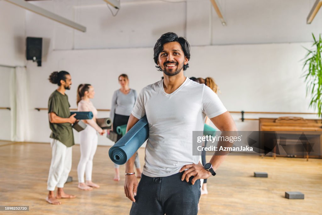Glücklicher erwachsener indischer Yogalehrer, der in der Kamera posiert und lächelt, während seine Kunden am Ende des Kurses im Hintergrund ein Gespräch führen