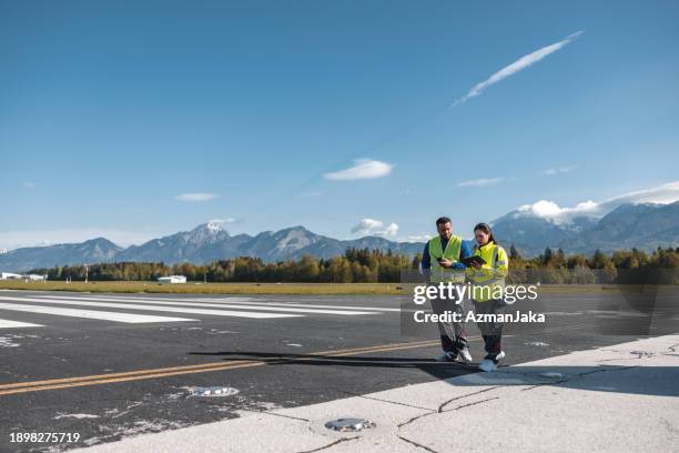 focused multi ethnic airport workers wearing yellow safety jackets while standing in the middle off an airstrip, they are using walkie talkie and electronic tablet device - airfield stock pictures, royalty-free photos & images