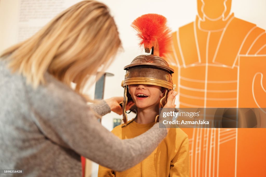 Lächelnder kaukasischer Junge probiert Eine Römische Galea Im Geschichtsmuseum, das Er Mit Seiner Attraktiven Blonden Mutter besucht
