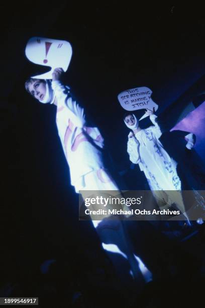 Fashion models wearing white outfits embellished with graffiti, holding speech bubbles by their heads, and bandages wrapped around their jaws, on the...