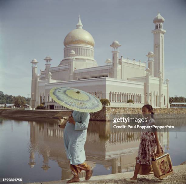 Two pedestrians walk beside a lagoon surrounding the Omar Ali Saifuddien Mosque in the city of Bandar Seri Begawan, capital of the British...