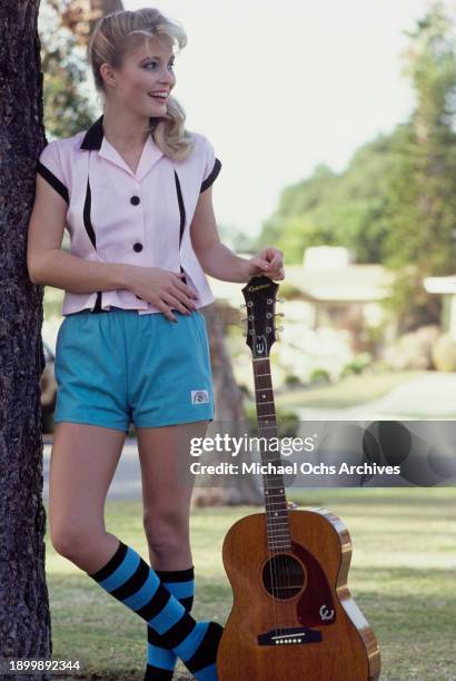 Fashion model modelling a sleeveless pale pink shirt with black trim, turquoise shorts, and blue-and-black striped socks, holding an acoustic guitar...