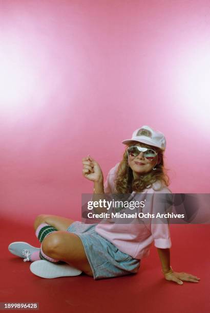 Fashion model modelling a pink sweatshirt, a grey skirt, a peaked cap, and sunglasses, sits on the studio floor during a studio fashion shoot,...