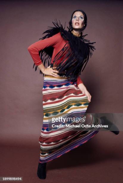 Fashion model wearing a red long-sleeved top beneath a black fringed vest with a long Aztec pattern skirt, during a studio fashion shoot, against a...