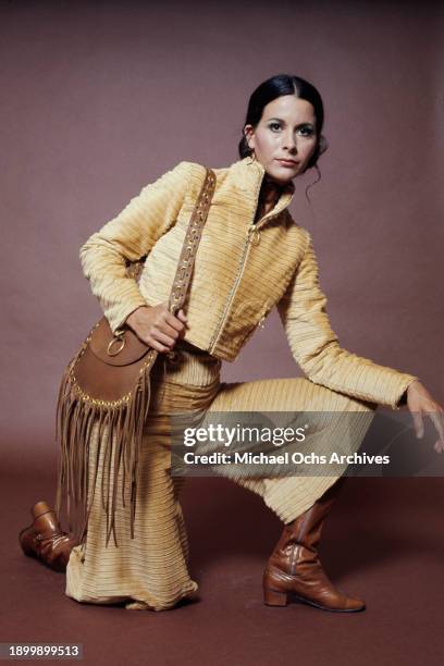 Fashion model wearing a gold-coloured trouser suit with wide-leg pedal pushers and brown boots, with a fringed brown purse, during a studio fashion...