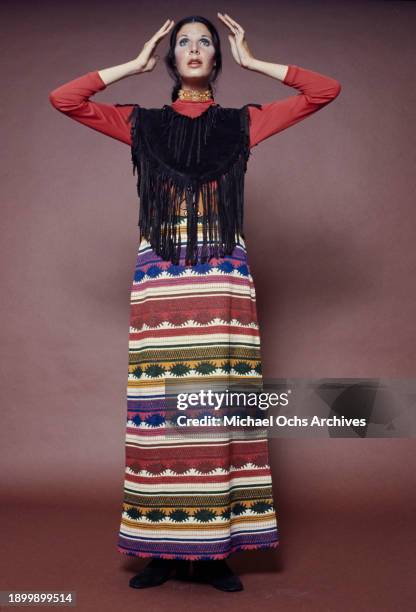 Fashion model wearing a red long-sleeved top beneath a black fringed vest with a long Aztec pattern skirt, holding her hands on either side of her...