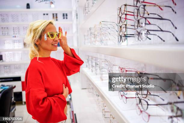 smiling young woman trying on glasses on mirror in optician - visual merchandise stock pictures, royalty-free photos & images