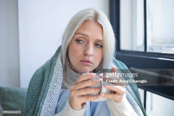 sad woman sits on windowsill looking through window at home. beautiful blonde lady with cup in her hands looking thoughtful, upset, lost in thoughts dreaming. - physical pressure stock pictures, royalty-free photos & images