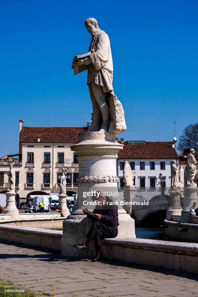 An old man sitting and reading at the foot of a statue on...
