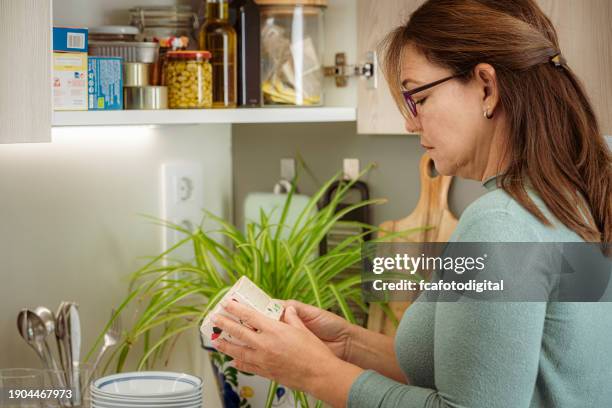 portrait of a woman in kitchen reading label of food package - nutrition label stock pictures, royalty-free photos & images