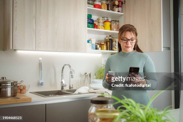 mujer hispana madura de pie en la cocina revisando la lista de comestibles en el teléfono inteligente - fechas de caducidad fotografías e imágenes de stock