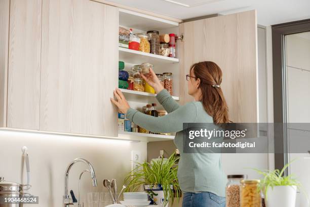 mujer toma un recipiente de vidrio de comida del gabinete superior de la cocina - organized kitchen pantry fotografías e imágenes de stock