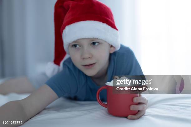 a boy in santa hat  lies on a bed on white linens and plays with a christmas tree toy holding cup christmas - big country breakfast stock-fotos und bilder