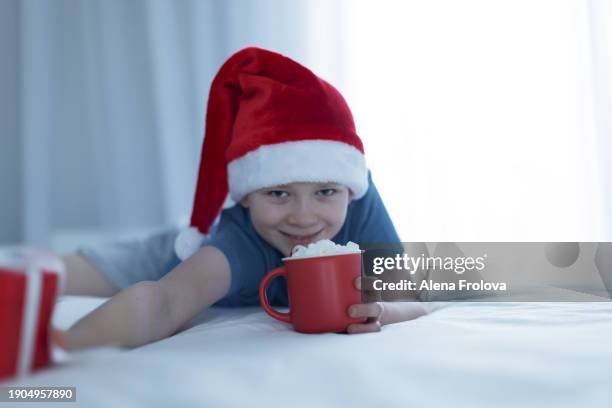 a boy in santa hat  lies on a bed on white linens and plays with a christmas tree toy holding cup christmas - big country breakfast stock-fotos und bilder