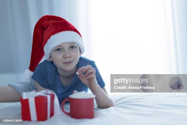 a boy in santa hat  lies on a bed on white linens and plays with a christmas tree toy holding cup christmas - big country breakfast stock-fotos und bilder