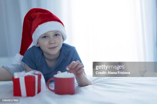 a boy in santa hat  lies on a bed on white linens and plays with a christmas tree toy holding cup christmas - big country breakfast stock-fotos und bilder