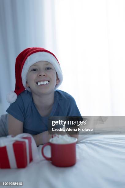 a boy in santa hat  lies on a bed on white linens and plays with a christmas tree toy holding cup christmas - big country breakfast stock pictures, royalty-free photos & images