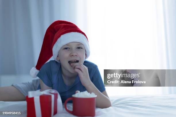 a boy in santa hat  lies on a bed on white linens and plays with a christmas tree toy holding cup christmas - big country breakfast stock pictures, royalty-free photos & images