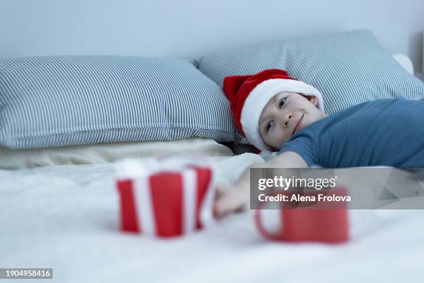 a boy in santa hat  lies on a bed on white linens and plays with a christmas tree toy holding cup christmas - big country breakfast stock pictures, royalty-free photos & images