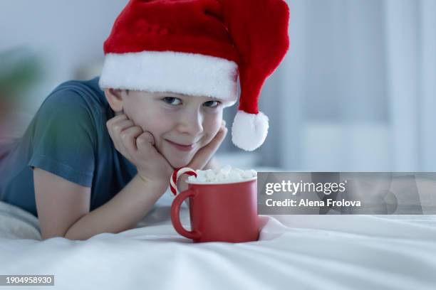 a boy in santa hat  lies on a bed on white linens and plays with a christmas tree toy holding cup christmas - big country breakfast stock-fotos und bilder