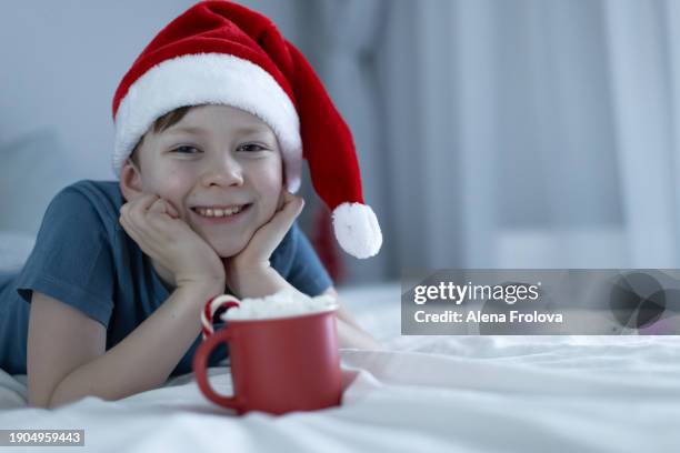 a boy in santa hat  lies on a bed on white linens and plays with a christmas tree toy holding cup christmas - big country breakfast stock-fotos und bilder