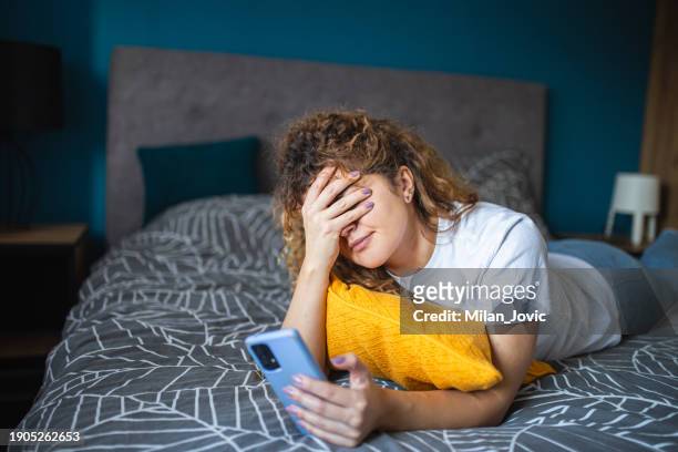 depressed woman lying on bed at home looking at mobile phone - female worried mobile imagens e fotografias de stock