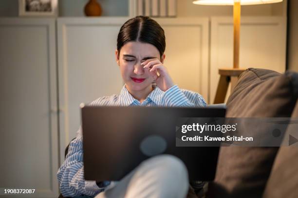 exhausted asian young woman hurt eye while using laptop in dark room. attractive girl sitting on sofa feeling visual fatigue and eye strain tired from overwork and massage dry irritable eye at home. - droger stockfoto's en -beelden
