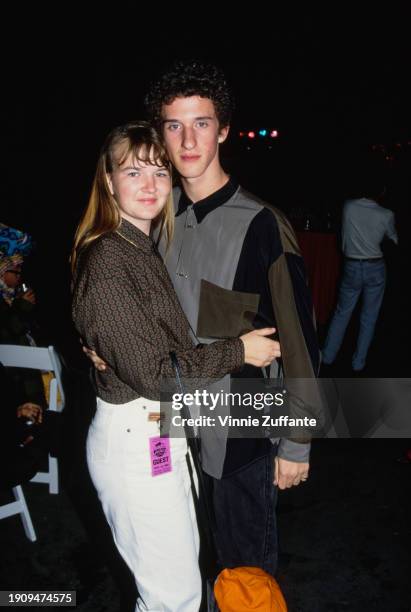 American actor and comedian Dustin Diamond, wearing a grey, black and olive green shirt, and embraced by a guest, who wears a patterned shirt and...