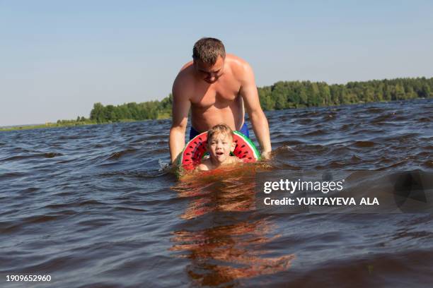 a small happy child boy has fun splashing water and swimming on a blue lake or river in an inflatable ring in the shape of a watermelon, observing safety precautions, together with his dad. father teaches child to swim - schwimmring stock-fotos und bilder