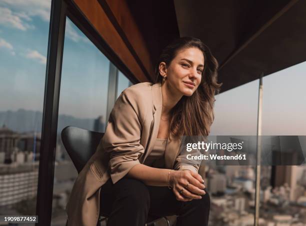 smiling businesswoman sitting on chair near window in office - witte blazer stockfoto's en -beelden