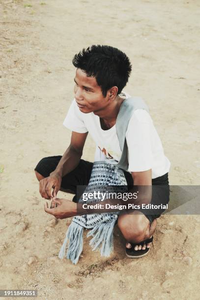 Boy Crouches On A Dirt Ground. Bagan, Myanmar.