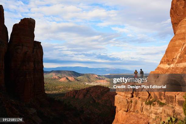 Two young hikers stand on red rock outcrop and enjoy a sweeping view of Sedona canyon and distant mountains. Sedona, Arizona, United States of...