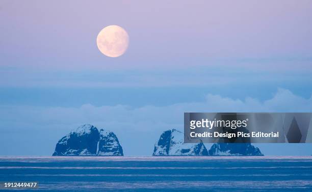 Full moon sets over two mountainous islands in the Gerlache Strait off the coast of the Antarctic peninsula. Antarctica.