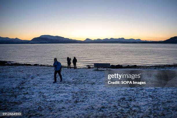 People walk along the shores of the Norwegian sea near the medieval church of Trondenes. Trondenes is a town in northern Norway, in the region of...