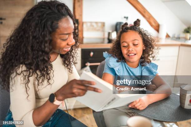 lovely diverse daughter and mother reading a book in a domestic dining room - family and happiness and diverse imagens e fotografias de stock