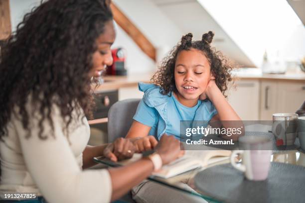 lovely diverse daughter and mother reading a book in a domestic dining room - family and happiness and diverse imagens e fotografias de stock