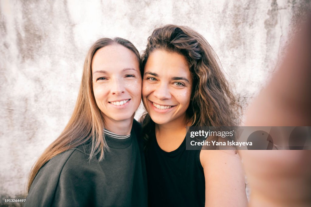A funny wide angle selfie of two diverse friends looking at camera, laughing. A fancy multicolor glitter on their young skin