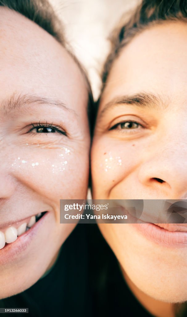 A funny wide angle selfie of two diverse friends looking at camera, laughing. A fancy multicolor glitter on their young skin