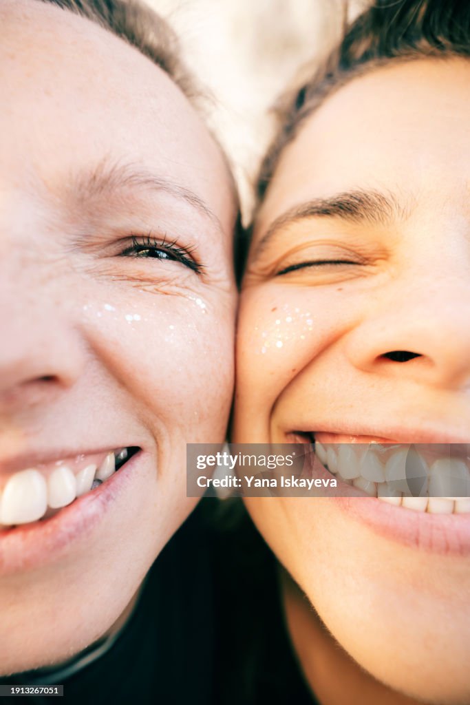 A funny wide angle selfie of two diverse friends looking at camera, laughing. A fancy multicolor glitter on their young skin