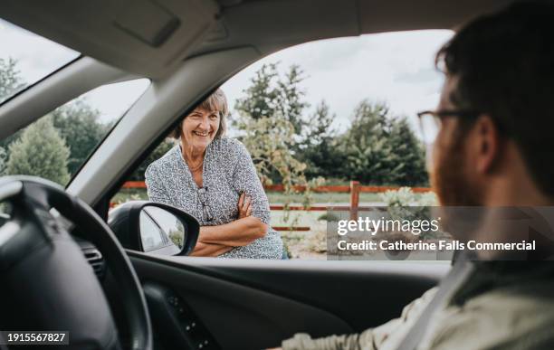 an older woman stands beside a car, looking through the window, and smiles in at the male driver - mom arms crossed stock pictures, royalty-free photos & images