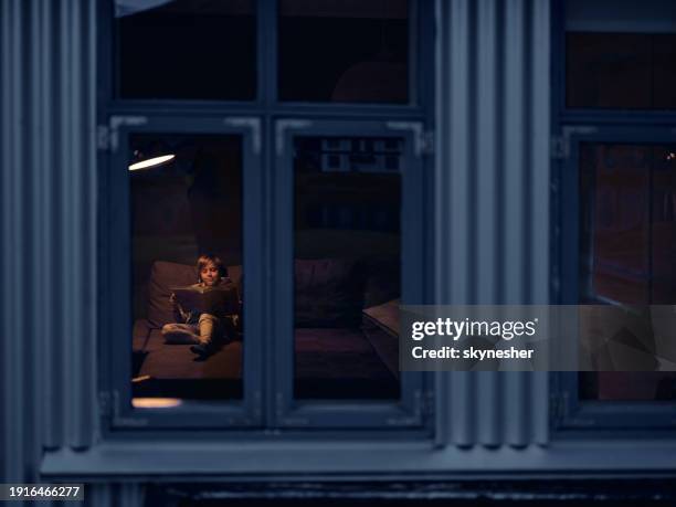 smiling boy reading a book while relaxing in the evening at home. - kid looking through window stock pictures, royalty-free photos & images