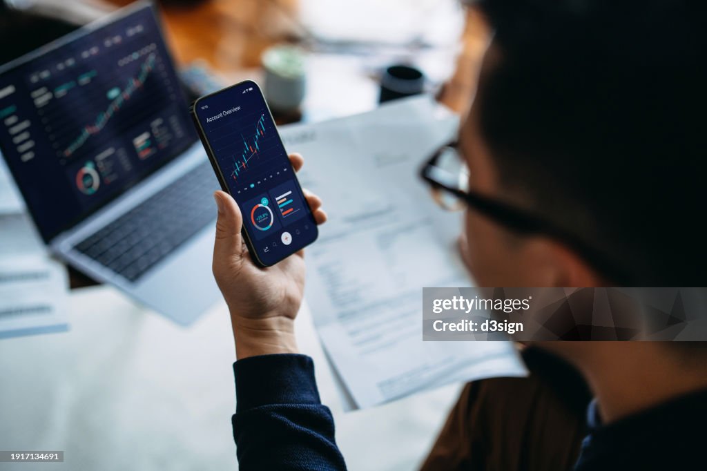 Young Asian man managing finance and investment online, analyzing stock market trades with smartphone and laptop. Making financial plans. Banking and finance, investment, financial trading, mobile banking with technology concept