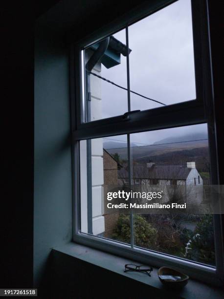 view through the window of a dark room, with houses under a cloudy gray sky - window frame stock pictures, royalty-free photos & images